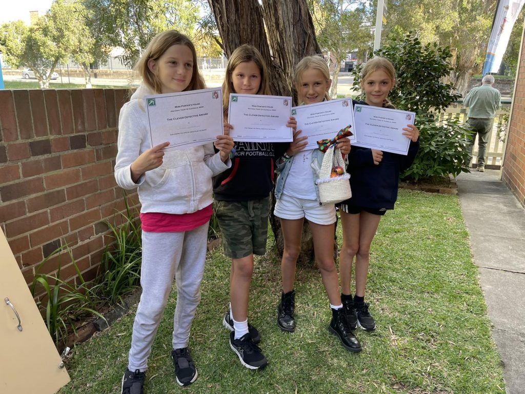 Four children form a line in front of a red brick wall, under the shade of an old tree. Children hold white participation certificates.
