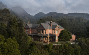 An imposing brick building with peaked grey roof surrounded by trees and wispy clouds due to the elevation.