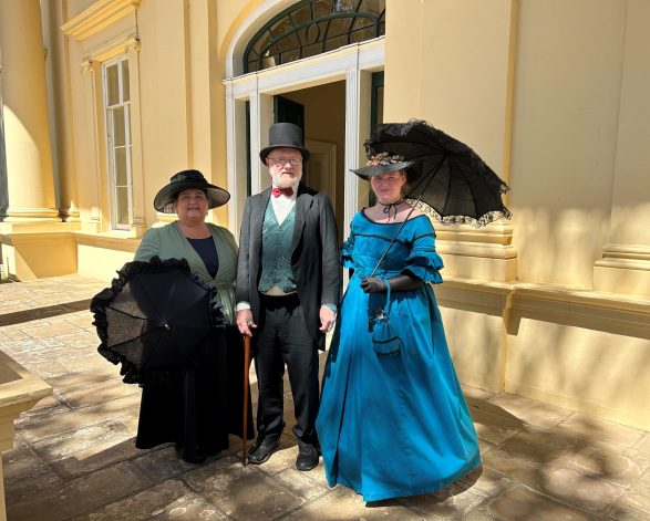 Two women in period costume flank a gentleman also in period costume They pose infront of an ornate door of a georgian house