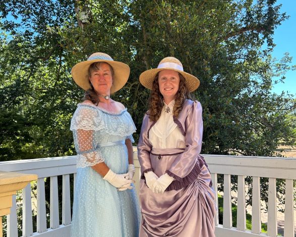 Two women pose elegantly against a white Georgian railing while dressed in period costume