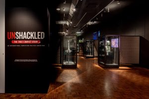 A dark museum room, most prominently a black wall with UNSHACKLED displayed. The floor is warm wood and showcases can be seen in the background.