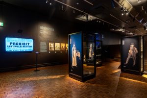 A dark museum room with brightly lit screens, tall showcases in dark colours and images of faces on the walls.